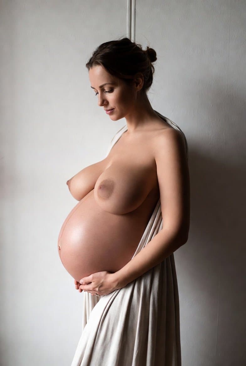 A pregnant woman poses against a plain wall, emphasizing her figure in a soft-lit photograph.